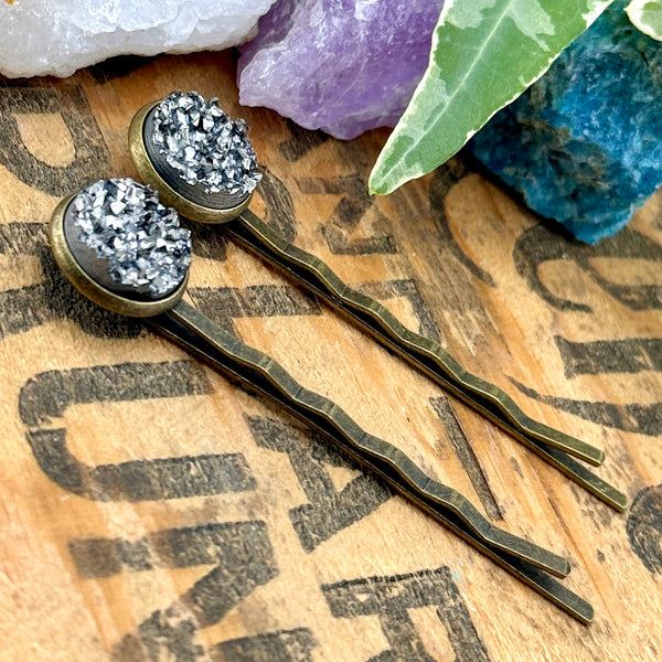 All Up In The Hair | Online Accessory Boutique Located in Mooresville, NC | Side view of two Pewter Druzy Bobby Pins laying on a wood background with black lettering. There are crystals and ivy leaves at the top of the image.
