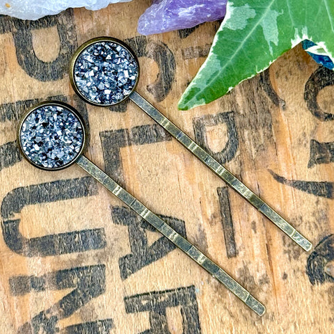 All Up In The Hair | Online Accessory Boutique Located in Mooresville, NC | Two metallic gray druzy bobby pins laying on a wood background with black lettering. There are crystals and ivy leaves at the top of the image.