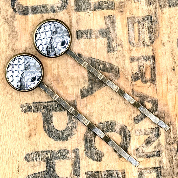 All Up In The Hair | Online Accessory Boutique Located in Mooresville, NC | Two Pewter Mermaid Bobby Pins laying on a wood background with black lettering.