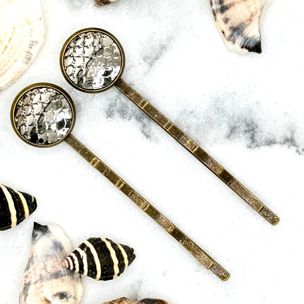 All Up In The Hair | Online Accessory Boutique Located in Mooresville, NC | Two Pewter Mermaid Bobby Pins laying on a white marble background surrounded by shells.