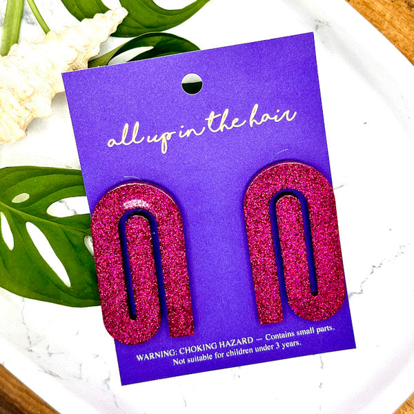 All Up In The Hair | Online Accessory Boutique Located in Mooresville, NC | Two bright pink paperclip earrings on an indigo backer card. The card is laying on a white background. Behind the card is some monstera leaves and a shell.