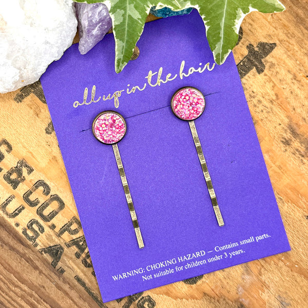 All Up In The Hair | Online Accessory Boutique Located in Mooresville, NC | Two Pink Spineal Druzy Bobby Pins on an indigo colored, All Up In The Hair branded packaging card. The card is laying on a wood background with black lettering. There are crystals and ivy leaves at the top of the image.