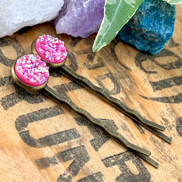 All Up In The Hair | Online Accessory Boutique Located in Mooresville, NC | Side view of two Pink Spineal Druzy Bobby Pins on a wood background with black lettering. There are crystals and ivy leaves at the top of the image.