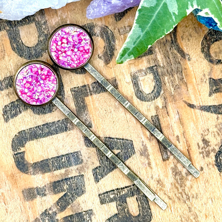 All Up In The Hair | Online Accessory Boutique Located in Mooresville, NC | Two Pink Spineal Druzy Bobby Pins laying diagonally on a grey background, surrounded by colorful glitter.