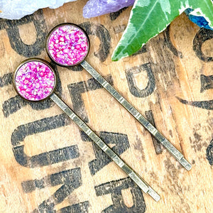All Up In The Hair | Online Accessory Boutique Located in Mooresville, NC | Two bright pink druzy bobby pins laying on a wood background with black lettering. There are crystals and ivy leaves at the top of the image.