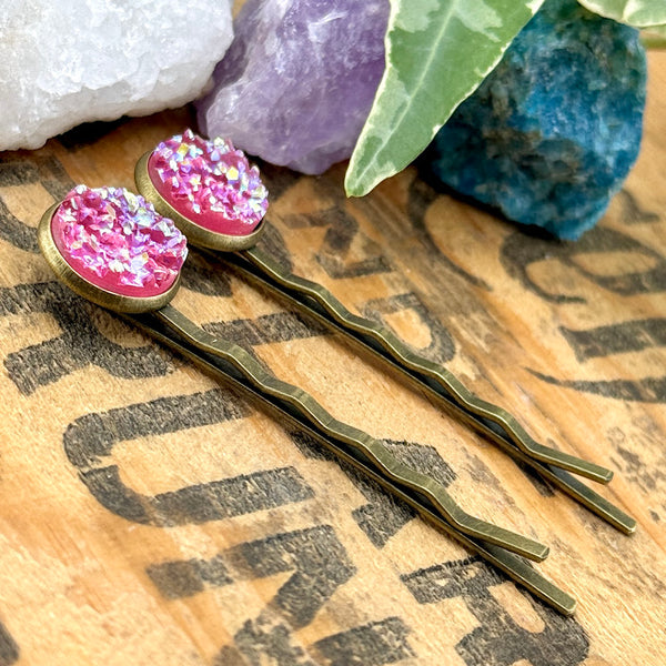 All Up In The Hair | Online Accessory Boutique Located in Mooresville, NC | Side view of our Pink Topaz Druzy Bobby Pins laying on a wood background with black lettering. There are ivy leaves and crystals at the top of the image.