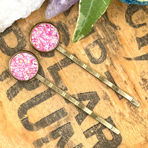 All Up In The Hair | Online Accessory Boutique Located in Mooresville, NC | Two pink druzy bobby pins laying diagonally on a grey background, surrounded by colorful glitter.