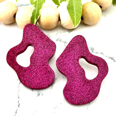 All Up In The Hair | Online Accessory Boutique Located in Mooresville, NC | Two Pink Wave Earrings on a white marble background. There is a wood bead garland and ivy leaves behind the earrings.