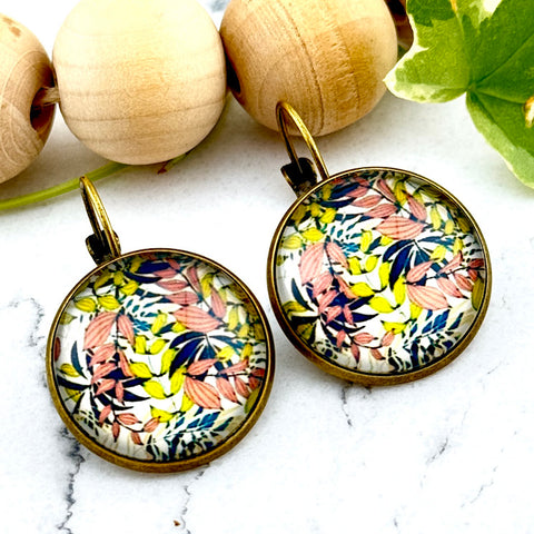 All Up In The Hair | Online Accessory Boutique Located in Mooresville, NC | Two round earrings with pink, blue and yellow leaves on a white marble background. Behind the earrings is a wood bead garland and ivy leaves.