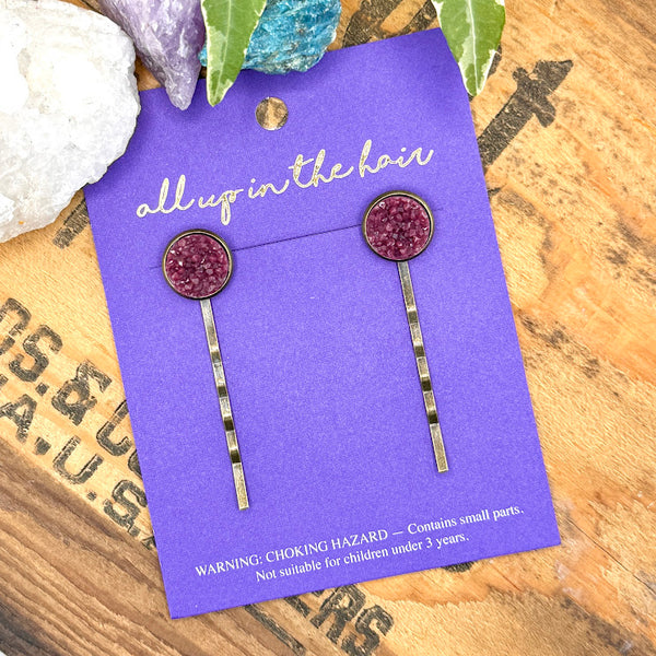 All Up In The Hair | Online Accessory Boutique Located in Mooresville, NC | Two Plum Druzy Bobby Pins on an indigo colored, All Up In The Hair branded packaging card. The card is laying on a wood background with black lettering. There are crystals and ivy leaves at the top of the image.