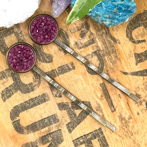 All Up In The Hair | Online Accessory Boutique Located in Mooresville, NC | Two plum colored druzy bobby pins laying diagonally on a wood background with black lettering. There are crystals and ivy leaves at the top of the image.