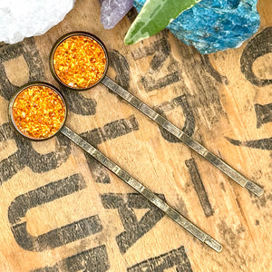 All Up In The Hair | Online Accessory Boutique Located in Mooresville, NC | Two orange druzy bobby pins laying on a wood background with black lettering. There are crystals and ivy leaves at the top of the image.