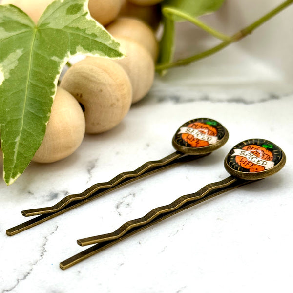 All Up In The Hair | Online Accessory Boutique Located in Mooresville, NC | Side view of two Pumpkin Spice Life Bobby Pins laying on a white marble background next to a wood bead garland and ivy leaves.
