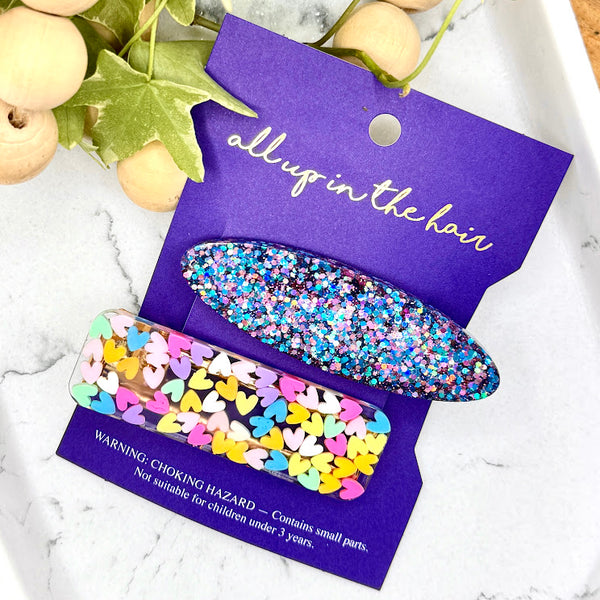 All Up In The Hair | Online Accessory Boutique Located in Mooresville, NC | All Up In The Hair branded card laying on a grey background surrounded by red glitter hearts. On the card is a pink and blue glitter oval barrette and a rectangular barrette made with pastel hearts.