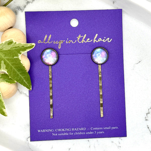 All Up In The Hair | Online Accessory Boutique Located in Mooresville, NC | Two Purple Galaxy Bobby Pins on an indigo colored, All Up In The Hair branded packaging card. The card is laying on a white marble background with a wood bead garland and ivy leaves.