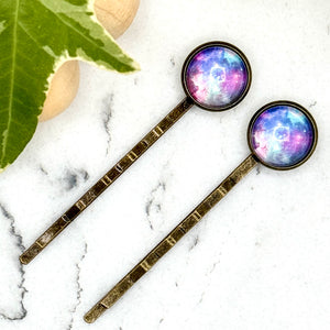 All Up In The Hair | Online Accessory Boutique Located in Mooresville, NC | Two Purple Galaxy Bobby Pins on a white marble background with a wood bead garland and ivy leaves.