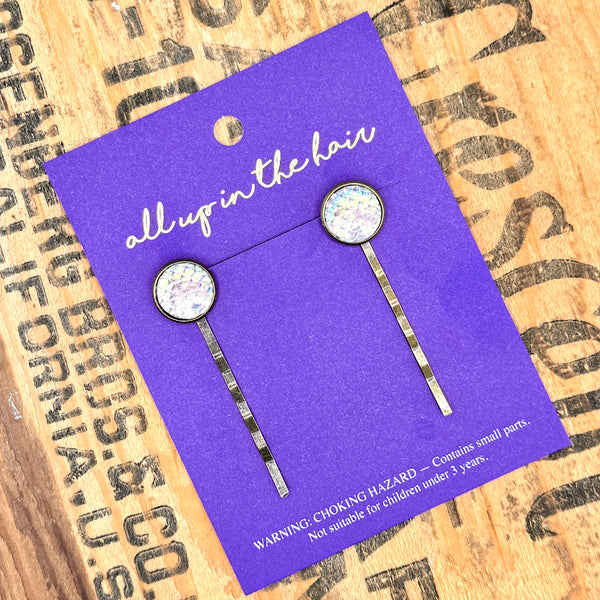 All Up In The Hair | Online Accessory Boutique Located in Mooresville, NC | Two Purple Mermaid Bobby Pins on an indigo colored, All Up In The Hair branded packaging. The card is laying on a wood background with black lettering.