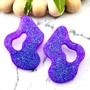 All Up In The Hair | Online Accessory Boutique Located in Mooresville, NC | Two Purple Wave Earrings on an white marble background. There is a wood bead garland and ivy leaves behind the earrings.
