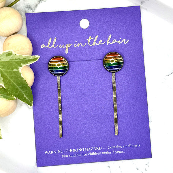All Up In The Hair | Online Accessory Boutique Located in Mooresville, NC | Two Rainbow Bobby Pins on an indigo colored, All Up In The Hair branded packaging card. The card is laying on a white marble background with a wood bead garland and ivy leaves.