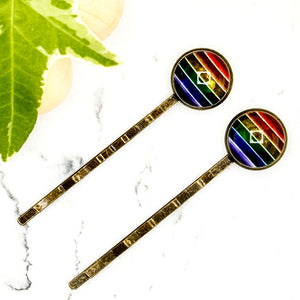 All Up In The Hair | Online Accessory Boutique Located in Mooresville, NC | Two Rainbow Bobby Pins laying on a white marble background with a wood bead garland and ivy leaves.