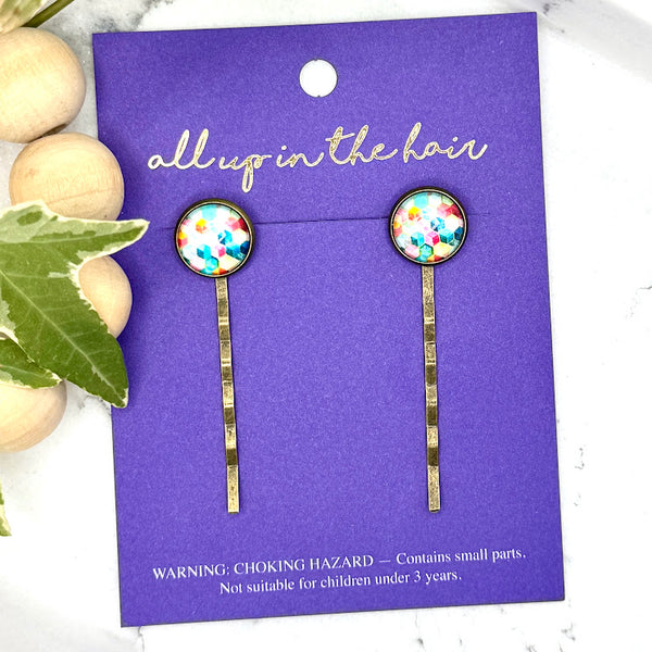 All Up In The Hair | Online Accessory Boutique Located in Mooresville, NC | Two Rainbow Geometric Bobby Pins on an indigo colored, All Up In The Hair branded packaging card. The card is laying on a white marble background with a wood bead garland and ivy leaves.