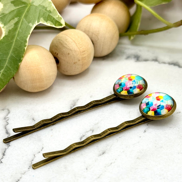 All Up In The Hair | Online Accessory Boutique Located in Mooresville, NC | Side view of two Rainbow Geometric Bobby Pins laying on a white marble background with a wood bead garland and ivy leaves.