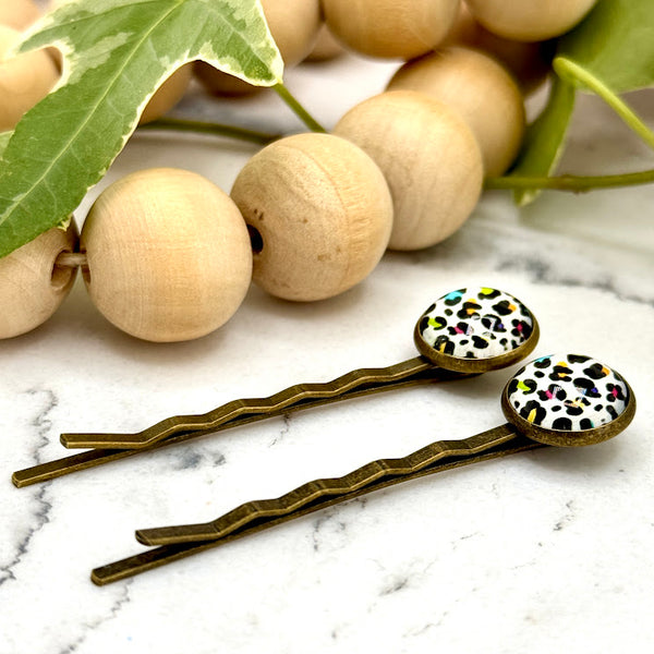 All Up In The Hair | Online Accessory Boutique Located in Mooresville, NC | Side view of two Rainbow Leopard Bobby Pins on a white marble background with a wood bead garland and ivy leaves.