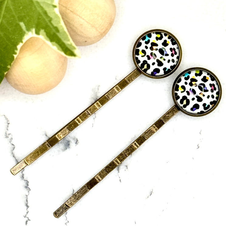 All Up In The Hair | Online Accessory Boutique Located in Mooresville, NC | Two Rainbow Leopard Bobby Pins laying on a white marble background with a wood bead garland and ivy leaves.