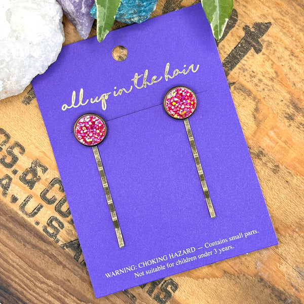 All Up In The Hair | Online Accessory Boutique Located in Mooresville, NC | Two Red Druzy Bobby Pins on an indigo colored, All Up In The Hair branded packaging card. The card is laying on a wood background with black lettering. There are crystals and ivy leaves at the top of the image.