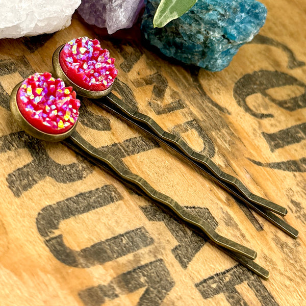 All Up In The Hair | Online Accessory Boutique Located in Mooresville, NC | Two Red Druzy Bobby Pins on a wood background with black lettering. There are crystals and ivy leaves at the top of the image.