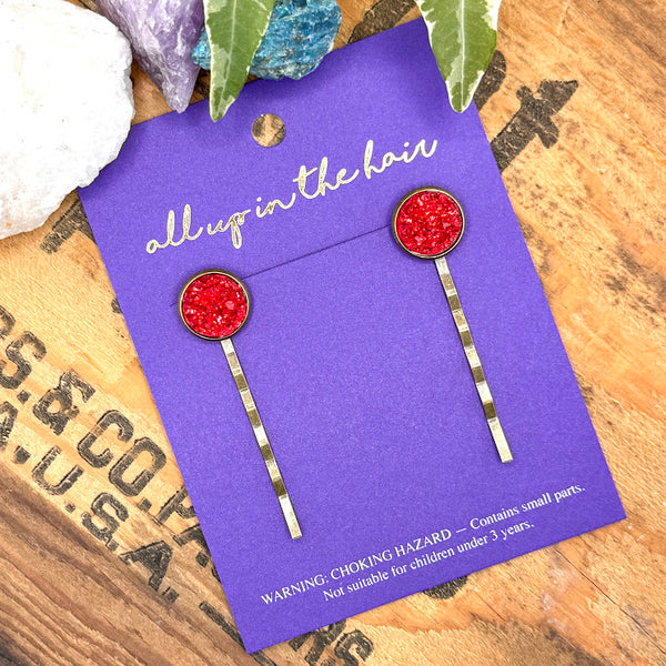 All Up In The Hair | Online Accessory Boutique Located in Mooresville, NC | Two Rhodochrosite Druzy Bobby Pins on an indigo colored, All Up In The Hair branded packaging card. The card is laying on a wood background with black lettering. There are ivy leaves and crystals at the top of the image.