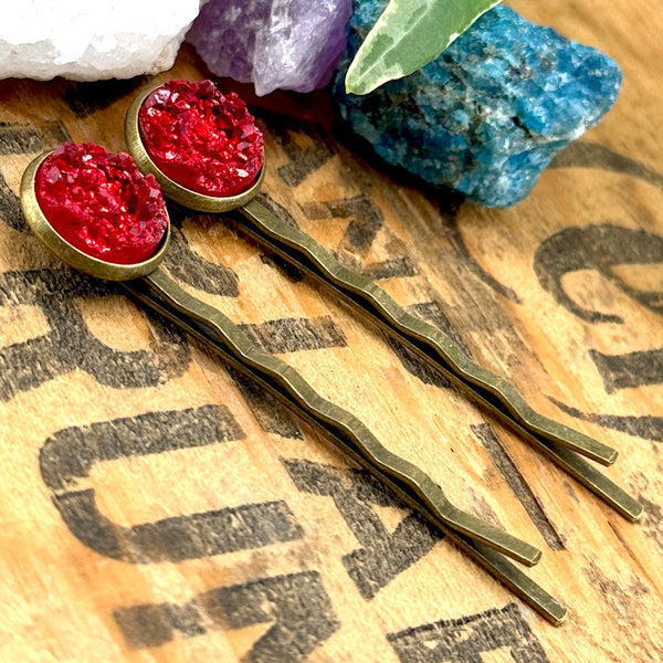 All Up In The Hair | Online Accessory Boutique Located in Mooresville, NC | Two Rhodochrosite Druzy Bobby Pins laying on a wood background with black lettering. There are crystals and ivy leaves at the top of the image.