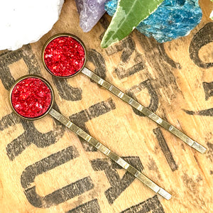All Up In The Hair | Online Accessory Boutique Located in Mooresville, NC | Two dark red druzy bobby pins laying diagonally on a grey background, surrounded by colorful glitter.