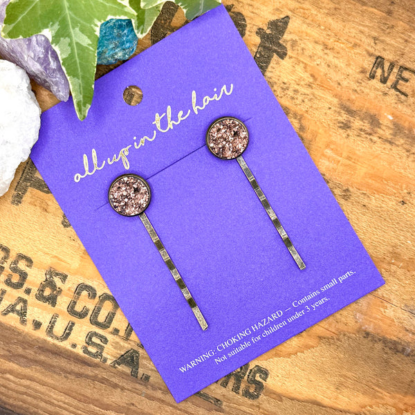 All Up In The Hair | Online Accessory Boutique Located in Mooresville, NC | Two Rose Gold Druzy Bobby Pins on an indigo colored, All Up In The Hair branded packaging card. The card is laying on a wood background with black lettering. There are crystals and ivy leaves at the top of the image.