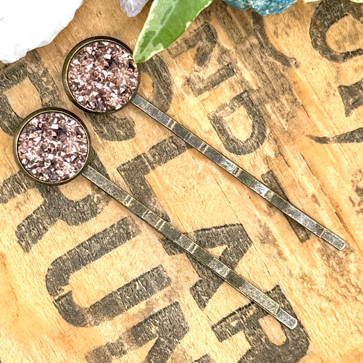 All Up In The Hair | Online Accessory Boutique Located in Mooresville, NC | Two rose gold druzy bobby pins laying diagonally on a wood background, surrounded by colorful glitter.