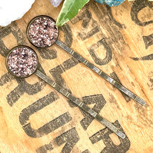 All Up In The Hair | Online Accessory Boutique Located in Mooresville, NC | Two Rose Gold Druzy Bobby Pins laying diagonally on a wood background with black lettering. There are crystals and ivy leaves at the top of the image.