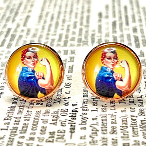 All Up In The Hair | Online Accessory Boutique Located in Mooresville, NC | Close up of two round earrings featuring Rosie the Riveter on a yellow background. The earrings are laying on a book page.