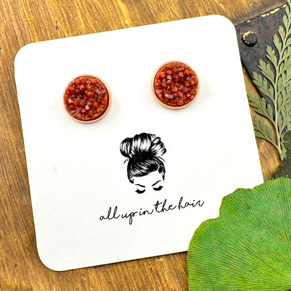 All Up In The Hair | Online Accessory Boutique Located in Mooresville, NC | Two rust colored druzies set in a rose gold earring setting. The earrings are on a white, All Up In The Hair branded packaging card. The card is laying on a wood background, alongside green leaves.