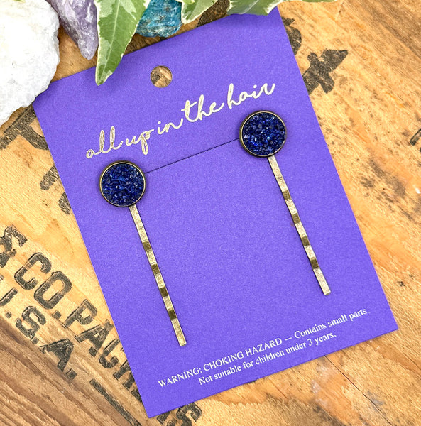 All Up In The Hair | Online Accessory Boutique Located in Mooresville, NC | Two Sapphire Druzy Bobby Pins on an indigo colored, All Up In The Hair branded packaging card. The card is laying on a wood background with black lettering. There are crystals and ivy leaves at the top of the image.