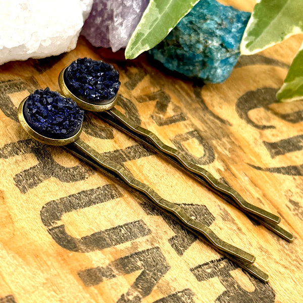 All Up In The Hair | Online Accessory Boutique Located in Mooresville, NC | Side view of our Sapphire Druzy Bobby Pins on a wood background with black lettering. There are crystals and ivy leaves at the top of the image.
