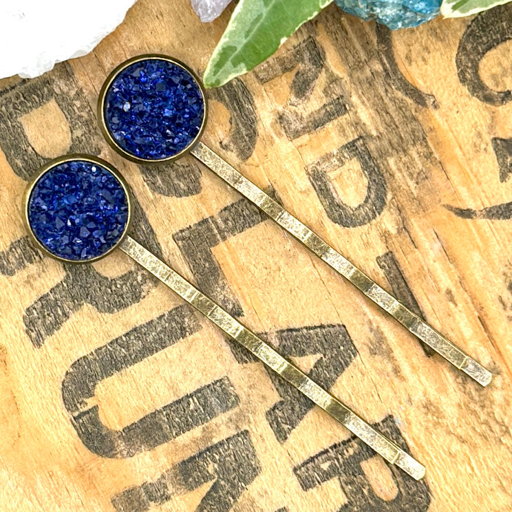 All Up In The Hair | Online Accessory Boutique Located in Mooresville, NC | Two dark blue druzy bobby pins are laying diagonally on a grey background, surrounded by colorful glitter.