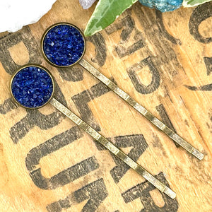 All Up In The Hair | Online Accessory Boutique Located in Mooresville, NC | Two dark blue druzy bobby pins on a wood background with black lettering. There are crystals and ivy leaves at the top of the image.