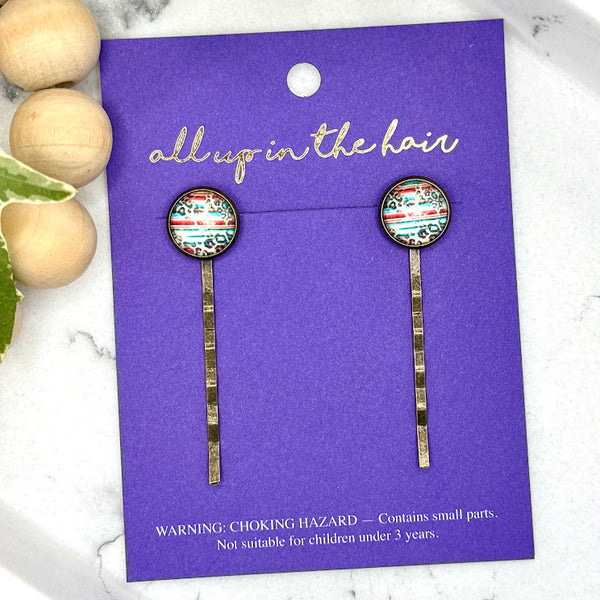 All Up In The Hair Wholesale | Online Accessory Boutique Located in Mooresville, NC | Two Serape Bobby Pins on an indigo backer card. Written above the earrings is "All Up In The Hair" in gold cursive. The card is laying on a book page.