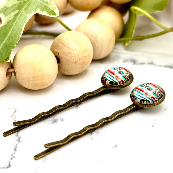 All Up In The Hair | Online Accessory Boutique Located in Mooresville, NC | Side view of two Serape Bobby Pins laying on a white marble background with a wood bead garland and ivy leaves.