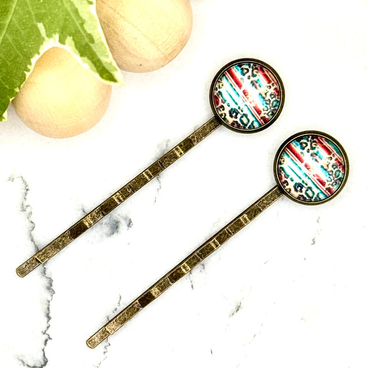All Up In The Hair | Online Accessory Boutique Located in Mooresville, NC | Two Serape and Leopard Print Bobby Pins on a white marble background with a wood bead garland and ivy leaves.