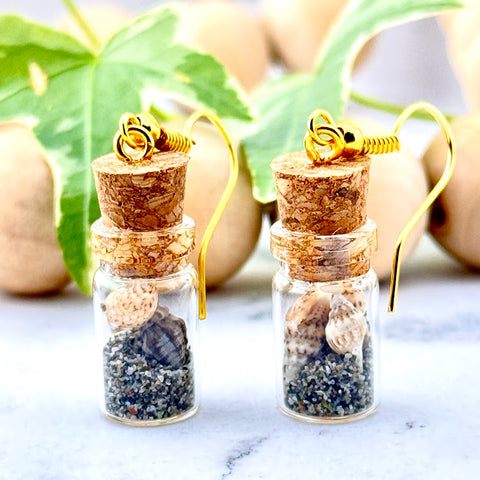 All Up In The Hair | Online Accessory Boutique Located in Mooresville, NC | Two tiny jars filled with dark sand and tiny shells on gold hooks. The earrings are sitting on a white marble background. Behind the earrings is a wood bead garland and ivy leaves.