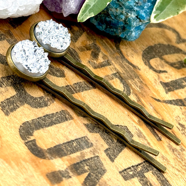 All Up In The Hair | Online Accessory Boutique Located in Mooresville, NC | Side view of two silver druzy bobby pins laying on a wood background with black lettering. There are crystals and ivy leaves at the top of the image.