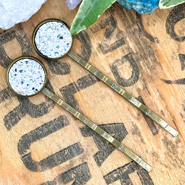 All Up In The Hair | Online Accessory Boutique Located in Mooresville, NC | Two silver druzy bobby pins laying diagonally on a grey background, surrounded by colorful glitter.