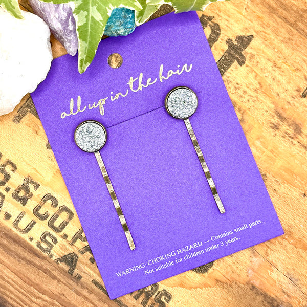 All Up In The Hair | Online Accessory Boutique Located in Mooresville, NC | Two Silver Glitter Druzy Bobby Pins on an indigo colored, All Up In The Hair branded packaging card. The card is laying on a wood background with black lettering. There are crystals and ivy leaves at the top of the image.