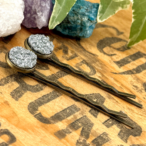 All Up In The Hair | Online Accessory Boutique Located in Mooresville, NC | Side view of our Silver Glitter Druzy Bobby Pins on an wood background with black lettering, There are crystals and ivy leaves at the top of the image.